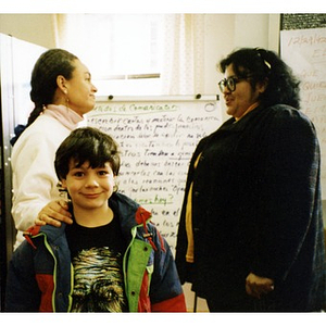 Boy smiling for the camera while two women stand behind him conversing.