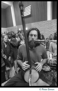 Krishna Das singing at a microphone, dotara in hand, during an appearance by Ram Dass at the College of Marin