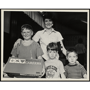 A woman poses with three boys, one of which holds the box for the board game "Careers: Parker Brothers Game of Fame, Fortune and Happiness"