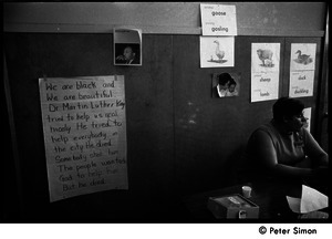 Teacher seated at a table at the Liberation School: in front of a poster honoring Martin Luther King: 'He tried to help everbody in the city. He died. Somebody shot him...'