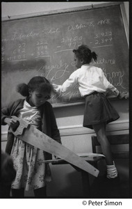 Girl using a saw while another girl writes on the blackboard, Liberation School, Boston, Mass.