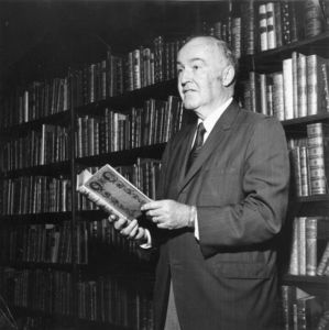 Poet Irving P. Zieman, reading from book, standing in front of bookcases at Suffolk University