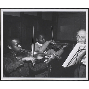 Two boys play violins while Henry Bernard, music director of the Boys' Clubs of Boston, turns the pages