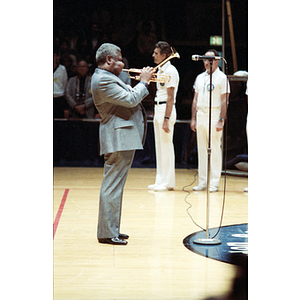 Man plays the trumpet into a microphone on a volleyball court, while two men stand behind him