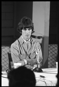 George Harrison seated in front of a microphone at a table, arms crossed, during a Beatles press conference