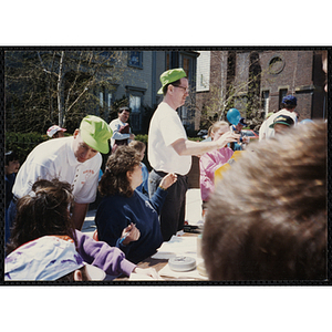 A male staff member standing with his right arm in front of him while another talks to a woman seated at a table at the Boys and Girls Clubs of Boston 100th Anniversary Celebration Street Fair