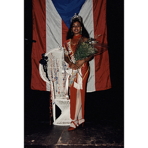 A woman poses in front of a Puerto Rican flag with a bouquet of flowers and wearing a crown