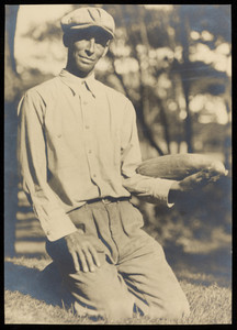 A man kneeling on the grass holding a cucumber or squash.
