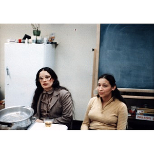 Two women sitting at a table in a room with a blackboard and a refrigerator.