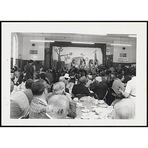 Guests, seated around tables, watch several girls performing on the stage at the South Boston Boys and Girls Clubhouse