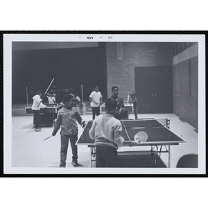 Boys play table tennis in a hall as another boy looks on