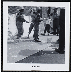 A man stands with a boy painting on plywood as other boys with paintbrushes look on during Tom Sawyer Day