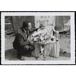 Miss South Boston, the winner of the Boys' Club Little Sister Contest, sits on her throne with a doll while a judge spreads her sash for a picture