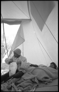 Inside of a strikers' tent: one woman huddled under blankets on the ground, another reaching into a bag