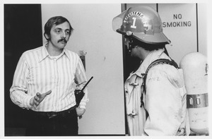 Bud Fay, Amherst Hockey Team Coach, standing indoors, talking with fireman