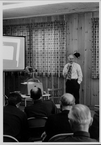 Ray Budde standing indoors, in front of group giving presentation