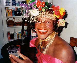 A Photograph of Marsha P. Johnson Wearing a Hot Pink Top While Lifting a Glass and Smiling