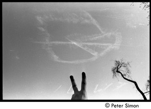 Protester flashing the v-sign beneath a skywriting peace symbol dissolving in the wind