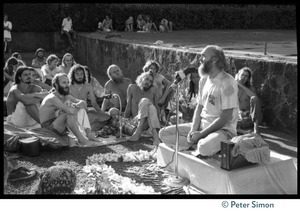 Ram Dass seated on a small platform, arms raised, during his appearance at Andrews Amphitheater, University of Hawaii