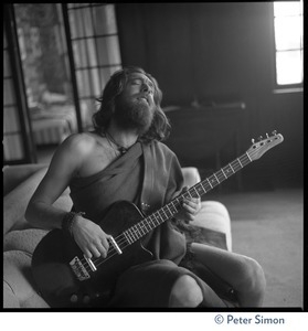 Krishna Das, playing bass guitar, sitting on a couch in the Vicente Street house