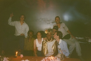 A Photograph From Marsha P. Johnson's Birthday Party Featuring Her Wearing a White and Gold Sequined Dress, Wearing a Floral and Pearl Headpiece, Smiling with Friends