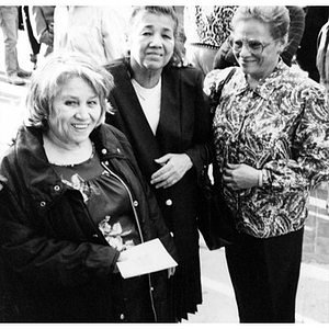 Three older women smiling for the camera as they stand outside in a crowd.