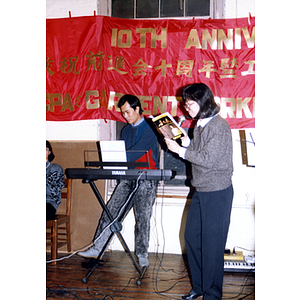 Facing left, a woman sings from a book while a man accompanies her on the keyboard at the Association's tenth anniversary celebration