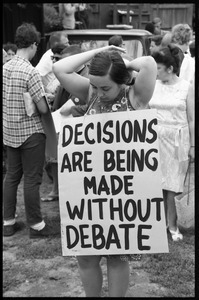Anti-war protester fixing her hair and wearing a sandwich board reading 'Decisions are being made without debate'