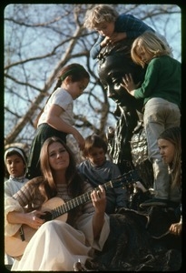 Judy Collins seated at the base of a statue, playing guitar for children