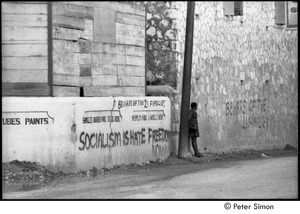 Woman leaning against a wall by graffiti reading, 'beware of the 21 families' and 'socialism is hate freedom now'