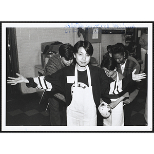 An Asian American boy looking at the camera with his arms spread as a staffer ties his apron behind his back during a Chelsea Boys and Girls Club event