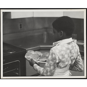 A boy carries a tray of buns in a kitchen