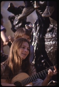 Judy Collins seated at the base of a statue, playing guitar for children