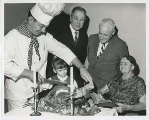 Chef carving turkey with a young client while Mr. Burrows, Mr. Place and an unidentified woman holding a bible look on