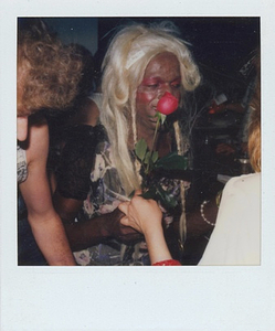 A Photograph of Marsha P. Johnson With Blonde Hair and Red Eyeshadow, Smelling a Rose