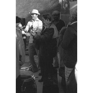 Onlookers watch as a male, Hispanic American plays a guitar (center); he is accompanied by a man playing maracas and wearing a Panama hat (on left)