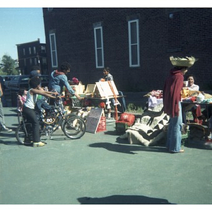 Two women sell decorative arts and crafts at tables at a Latino street festival, while adults and some children on bicycles look at the items