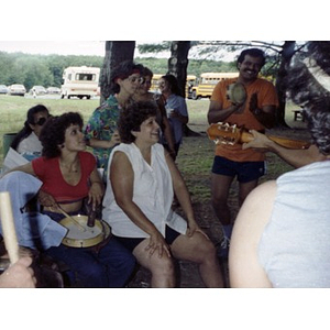 People play musical instruments while others listen and smile at a La Alianza staff picnic at an unidentified location in the woods