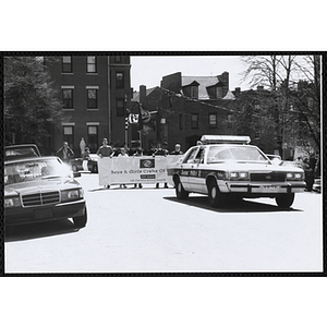 Several boys holding a large banner with the text "Boys and Girls Clubs of Boston 100 Years Of Celebrating Youth" and walking behind a police car during the parade