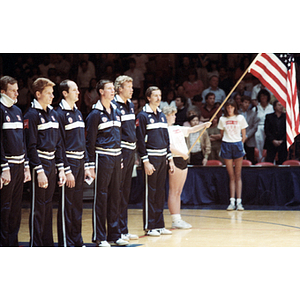 Members of the USA men's volleyball team stand in a line, while a woman at the end of the line holds up an American flag