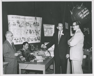 Two men watch a man at work at an eyeglass lens making display table