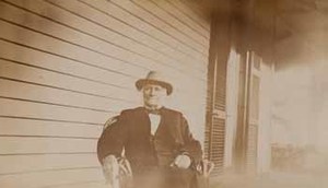 Sidney Bartlett seated in chair on porch, facing forward, with cigar