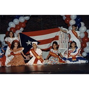 Two girls hold up a Puerto Rican flag as four girls kneel in front of it at the Festival Puertorriqueño