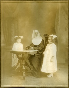 Girls instructed by a nun, possibly Easthampton, Mass.: full-length studio portrait with nun seated at a table, reading, possibly for first communion