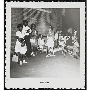 Participants' families sit and stand in front of a wall at a Boys' Club Little Sister Contest