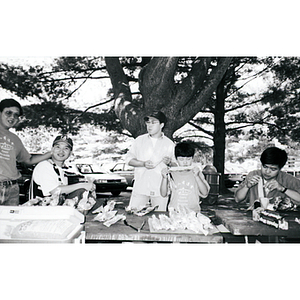 Young boy eats corn on the cob at a picnic table, with others standing and sitting nearby