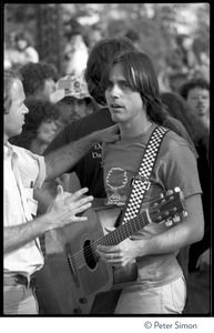 Jackson Browne, with acoustic guitar, talking to unidentified man at the No Nukes concert and protest, Washington, D.C.