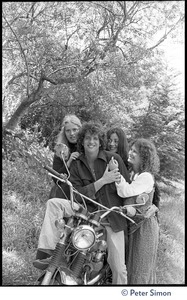 Peter Simon seated on a motorcycle, with Usha (Cathy Brown, right) and two other admiring women caressing him