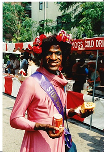 A Photograph of Marsha P. Johnson Holding a Sandwich and Soda, Wearing a Pink Dress and Purple “Stonewall” Sash