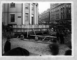 Construction at the corner of Park St., showing wood fence in front of a hole, with pedestrian and trolley traffic in the background